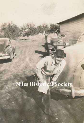 Phyllis' dad cleaning his boots after working in the barn. In the background is the Blott apple orchard which provided fruit for sale at the market. The car beside him is a 1941 Plymouth and older cars behind him are not identified.