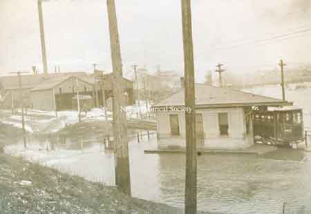 The frame Streetcar Waiting Station on Robbins Avenue, along Mosquito Creek on the north side of the street, just east of the overpass for the railroad. Note the streetcar barn in the background.