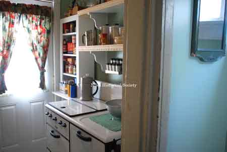 Detail showing kitchen area with stove, storage cabinet and back doorway with typical curtains.